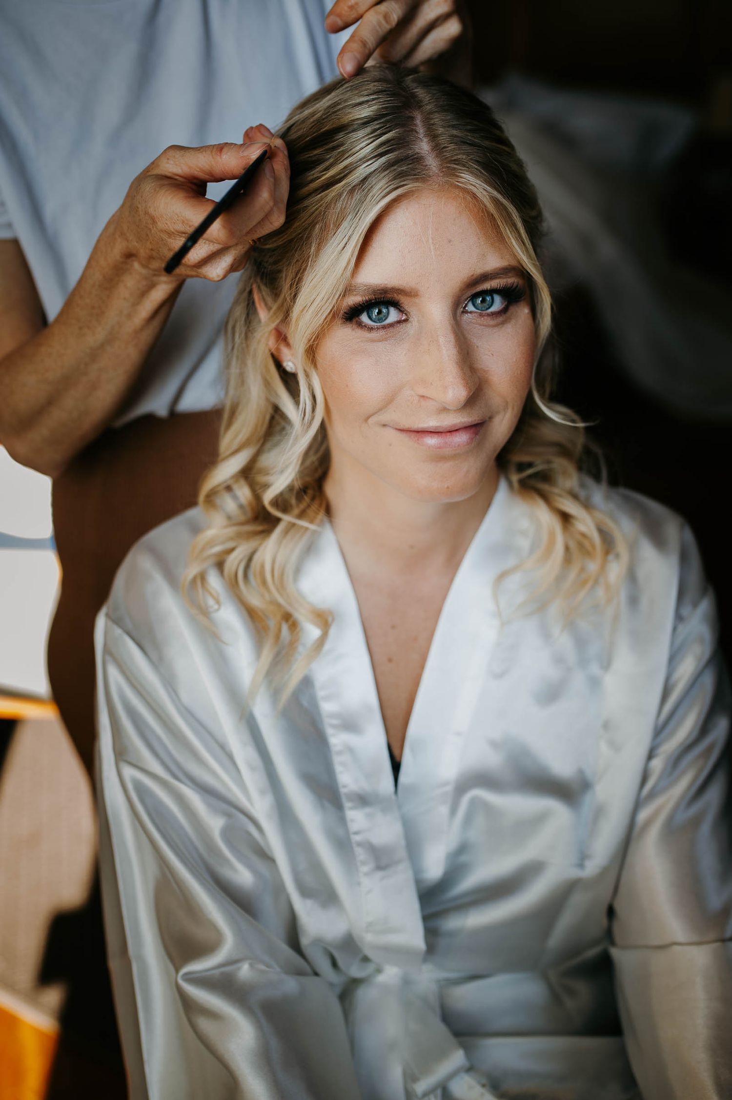 Blonde bride with blue eyes getting hair styled while wearing a white satin robe on wedding day.