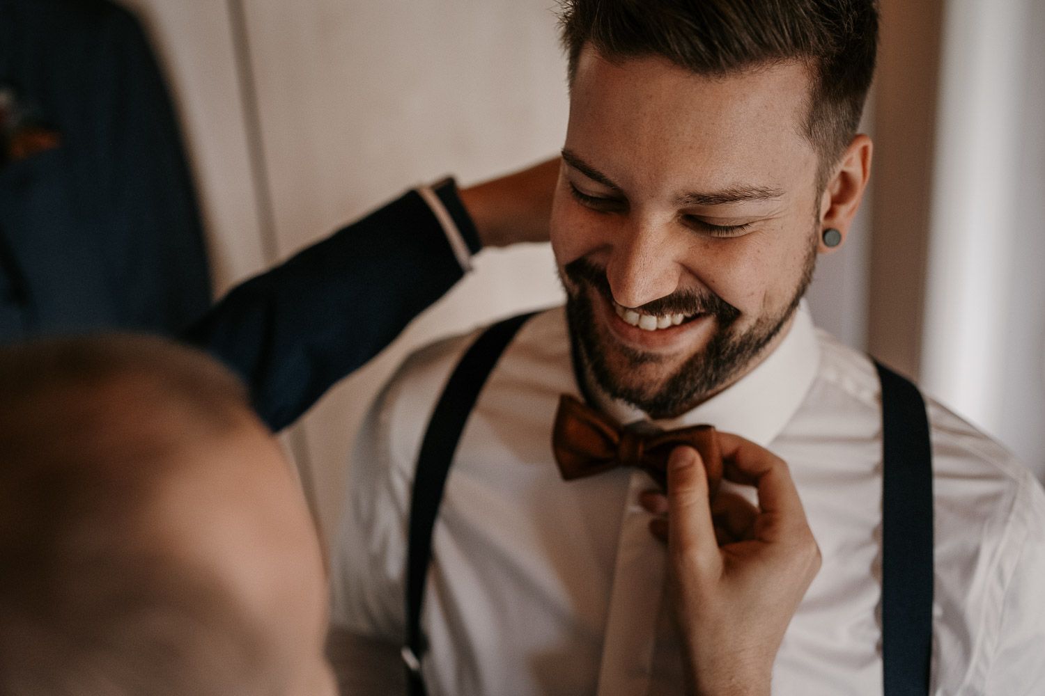 Smiling groom having a brown bow tie adjusted while wearing white shirt and blue suspenders.