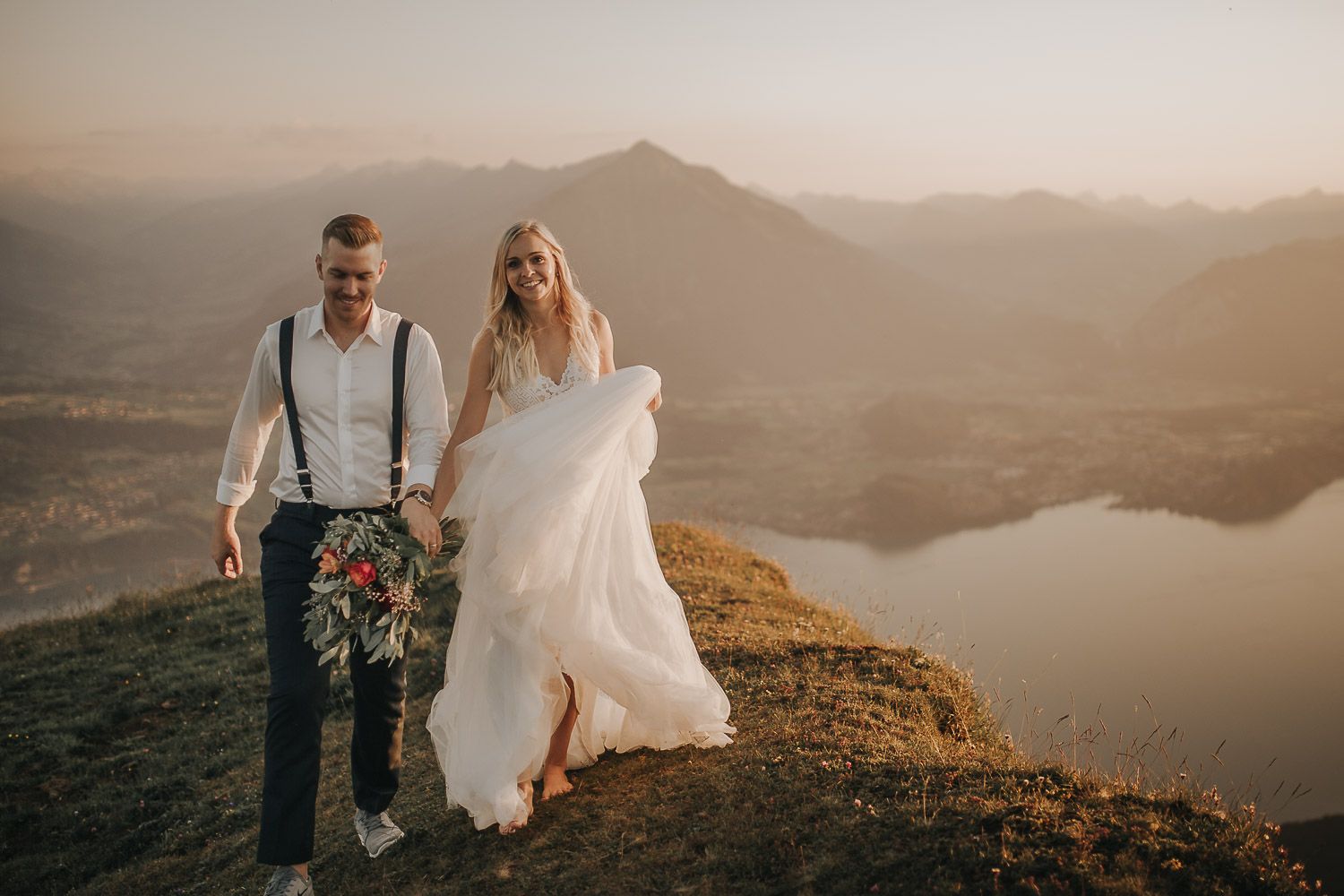 Adventurous wedding couple walking on mountain summit at sunset with stunning lake and Alpine panorama below.