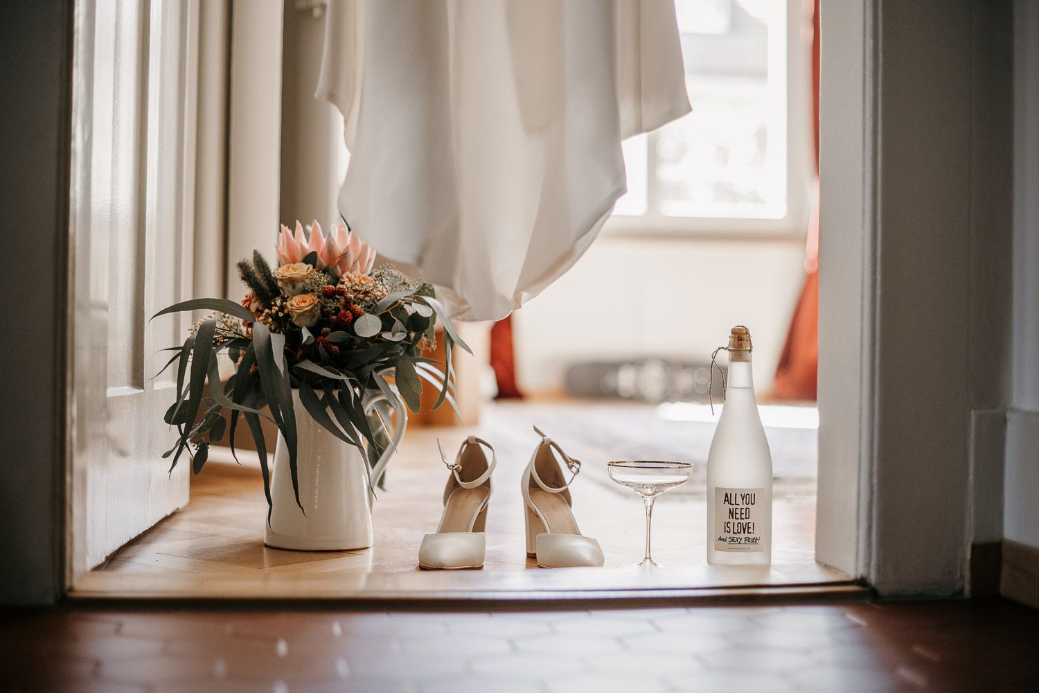 Bridal flat lay featuring a bouquet, white heels, champagne glass, and decorative bottle by a window.