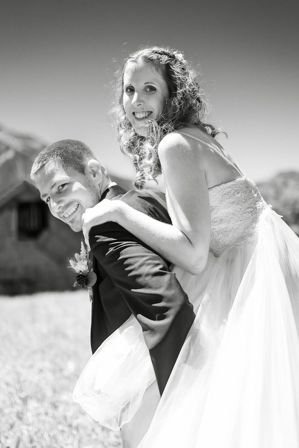 Happy bride piggybacks on smiling groom in black tuxedo outdoors on their wedding day, black and white photo.