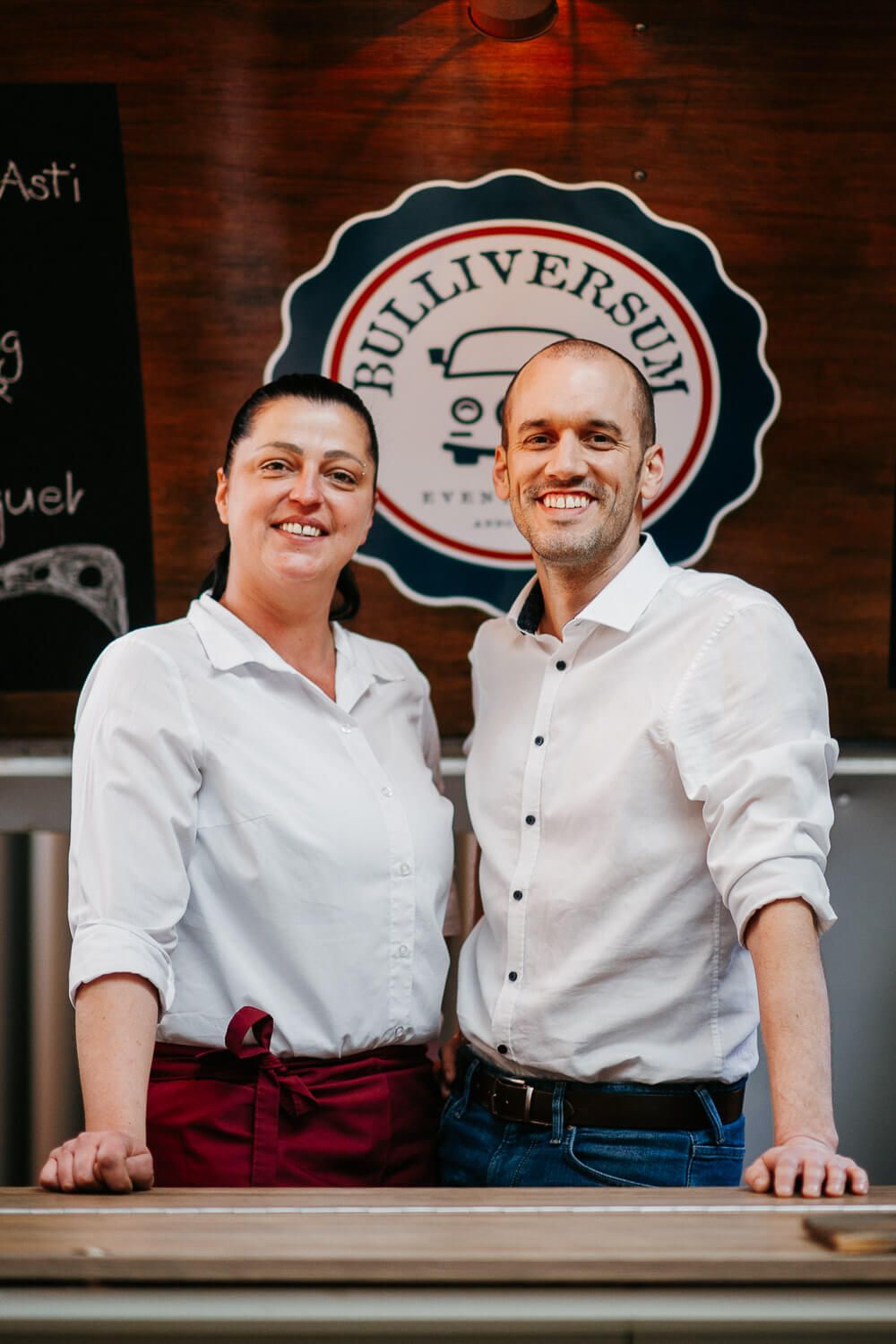 Two smiling restaurant staff in white shirts posing at counter in front of Bulliversum logo sign.