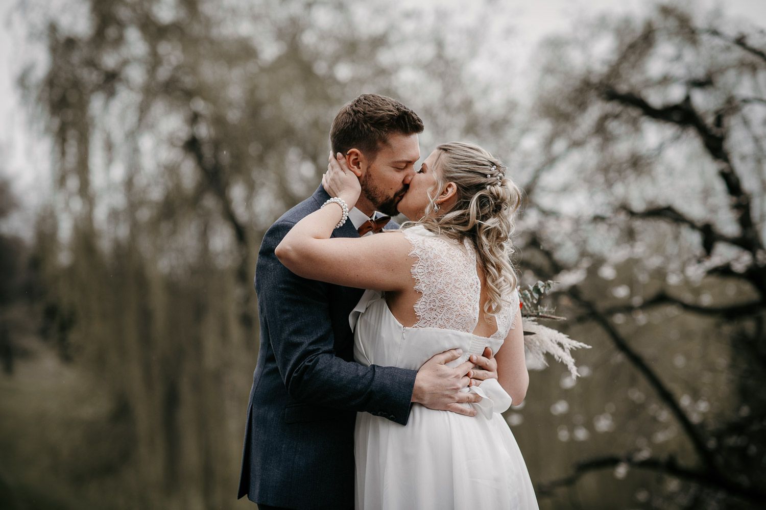 Bride and groom sharing a passionate kiss outdoors with blooming trees in the background.