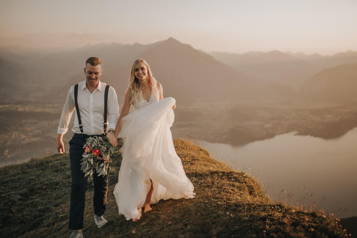 Bride and groom walking on mountain summit at golden hour with stunning lake and alpine backdrop.
