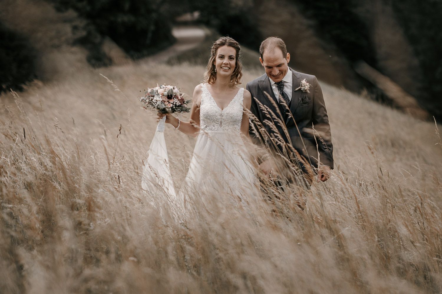 Happy bride and groom walking through golden wheat field, bride holding bouquet in a lace V-neck wedding gown.