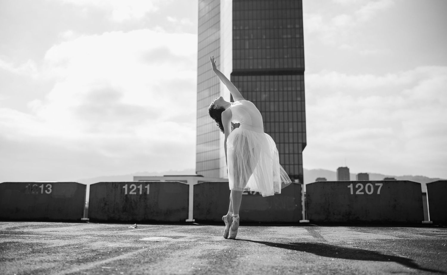 Black and white photo of ballerina dancing en pointe outdoors near urban skyscraper on rooftop.