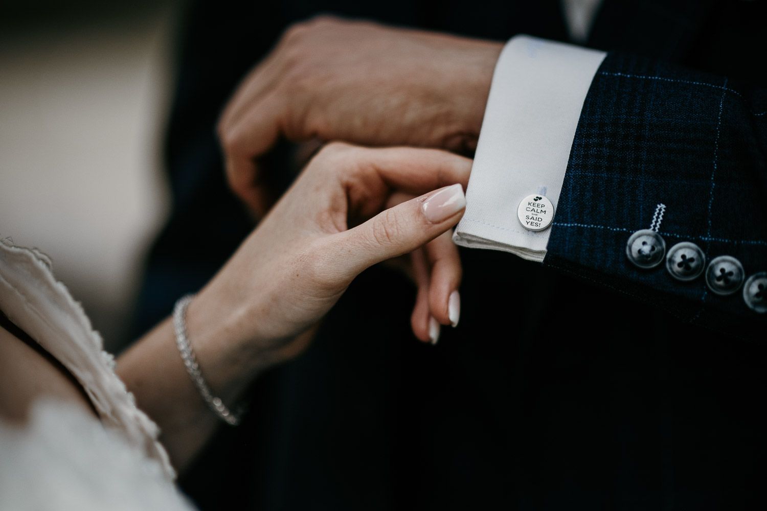 Bride fastening silver cufflinks on grooms navy plaid suit jacket sleeve before the wedding ceremony.