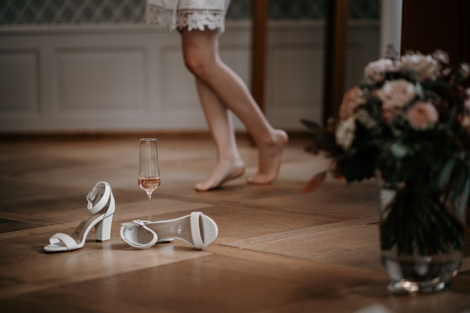 White bridal heels and a champagne glass on a wooden floor with a barefoot bride walking away.
