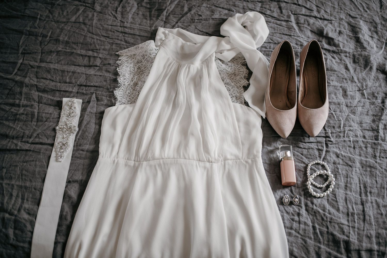 Bridal flat lay with white chiffon dress, blush heels, pearl bracelet, and perfume on gray linen.