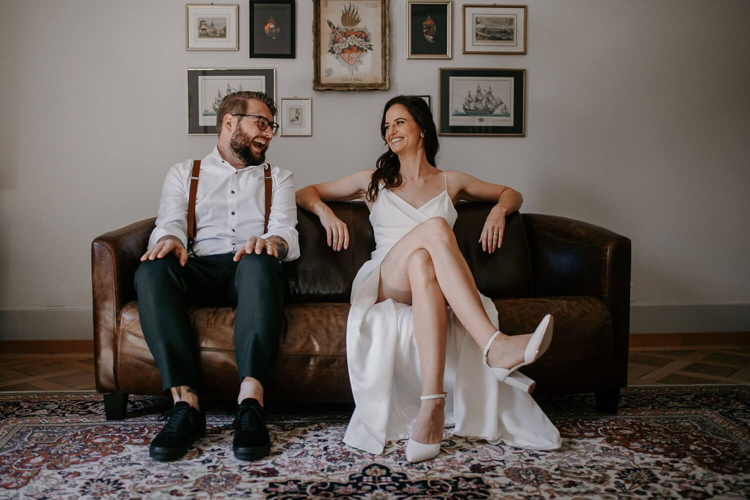 Bride and groom sitting on leather sofa laughing together in stylish indoor wedding portrait.