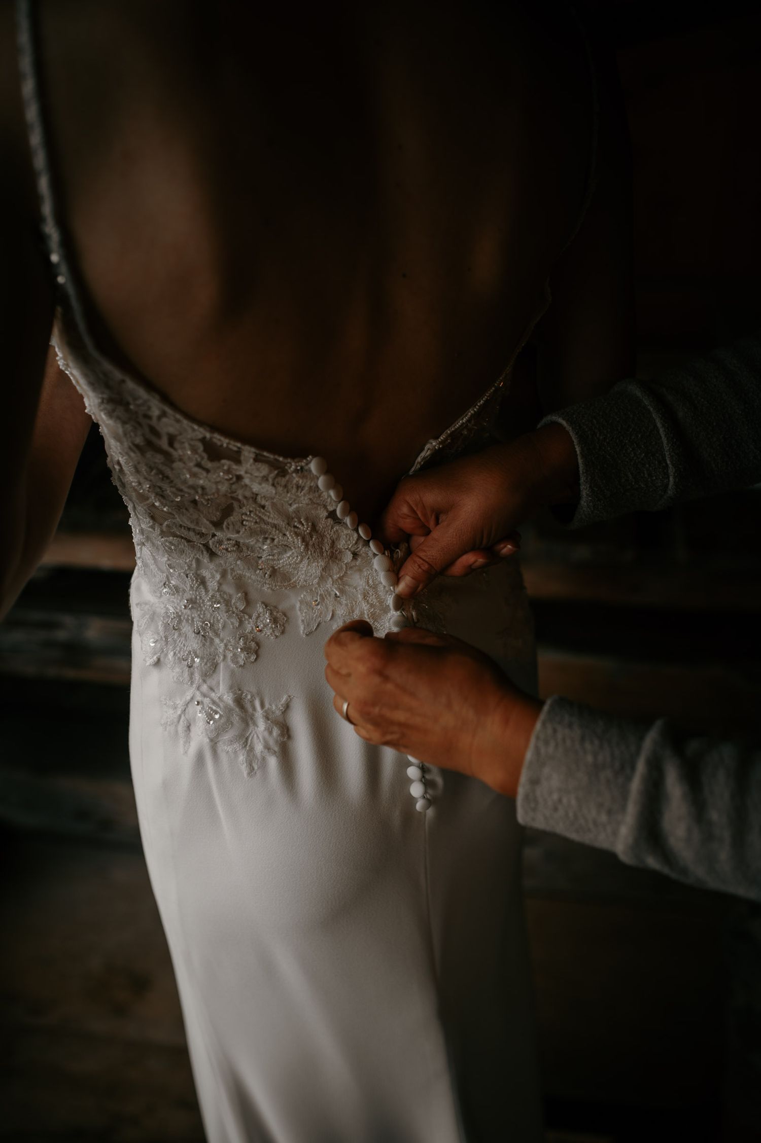 Hands fastening delicate buttons on the back of a lace wedding dress in moody lighting.
