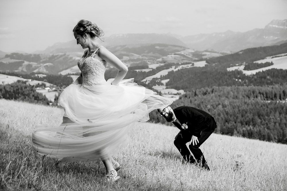 Black and white photo of bride twirling dress while groom bows on scenic hilltop with mountain valley backdrop.