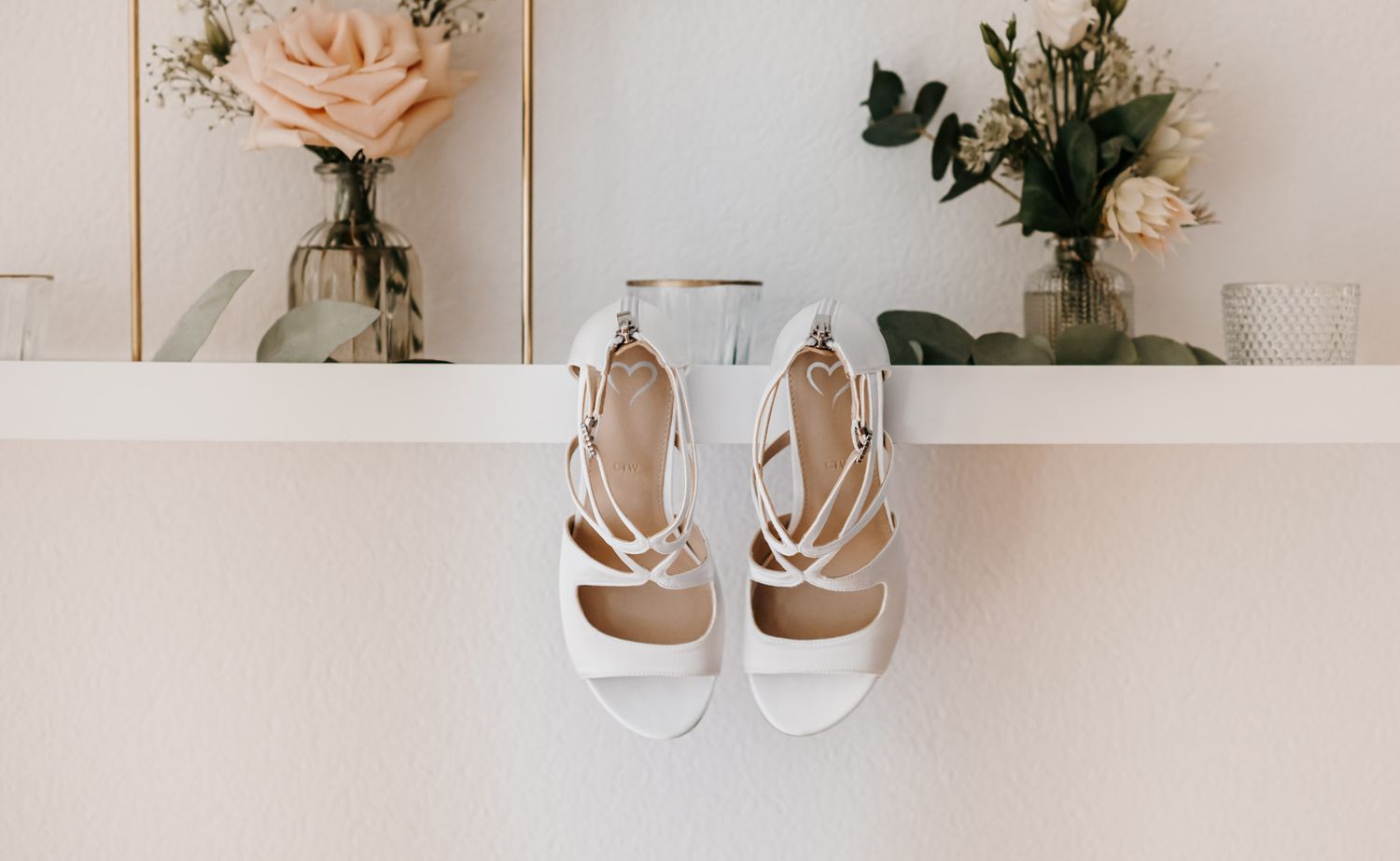White strappy bridal heels displayed on a shelf with floral arrangements in glass vases.