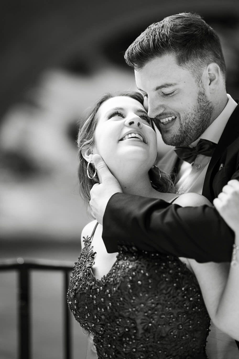 Groom tenderly embraces bride in elegant beaded gown, sharing a romantic moment in black and white portrait.