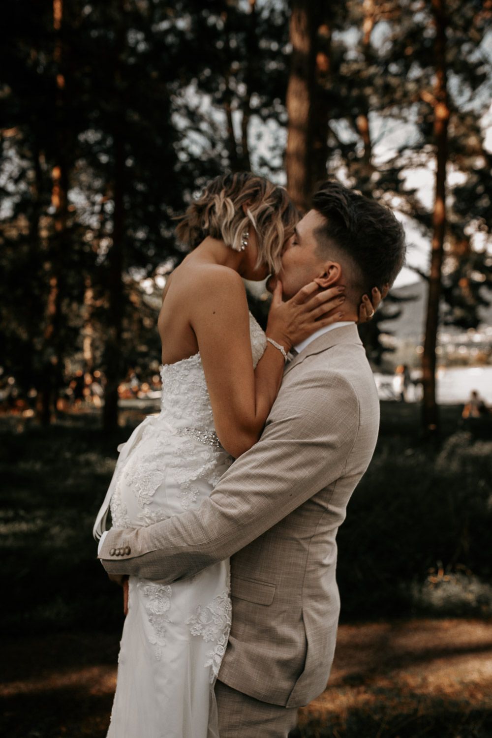 Romantic wedding couple sharing a tender kiss outdoors, bride in lace gown, groom in beige suit among trees.