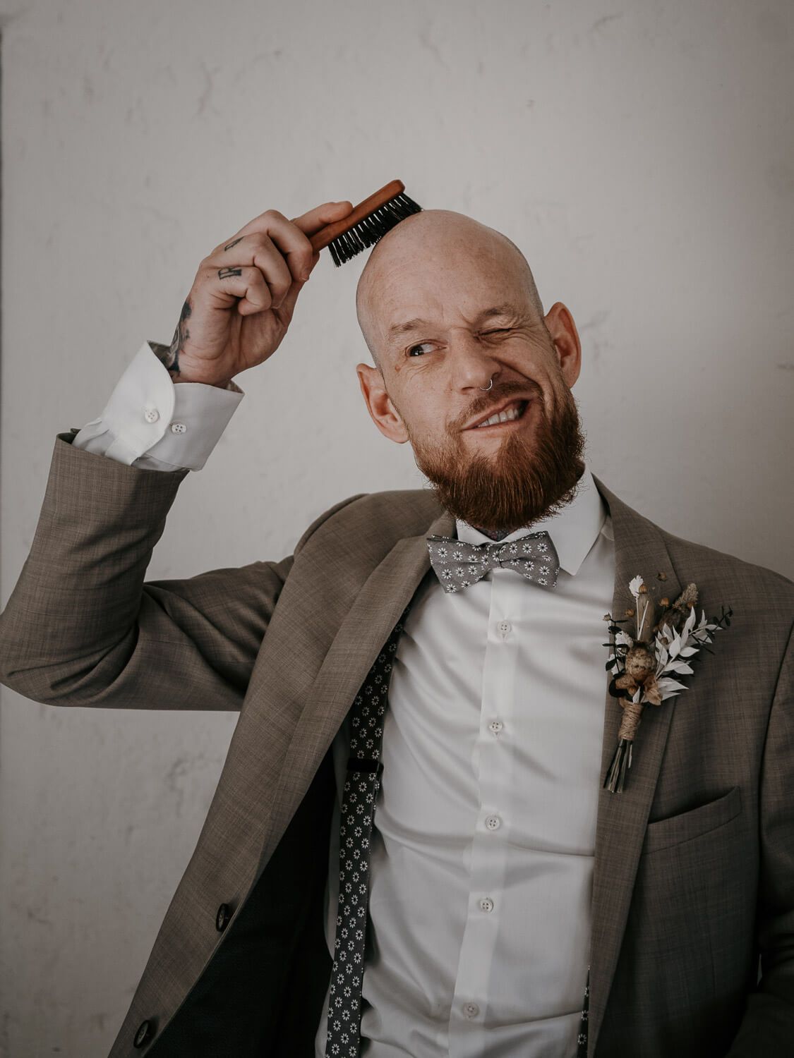 Bald bearded groom in gray suit humorously combing his head with a beard brush before wedding.