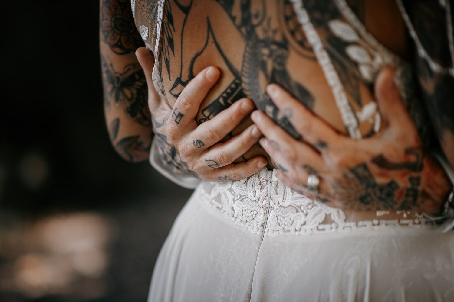Close-up detail of heavily tattooed hands clasped at the back of a bride in a white lace wedding dress.