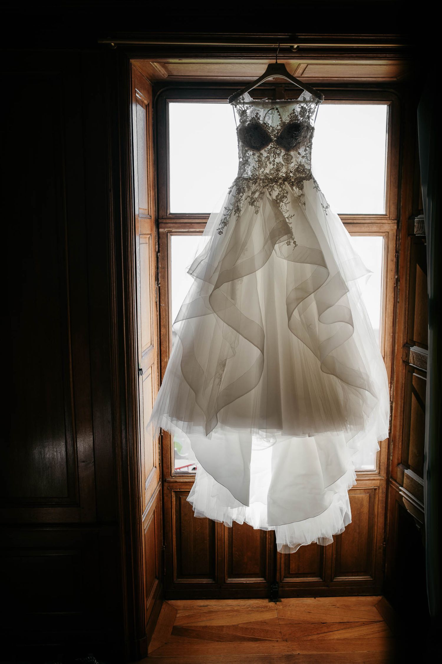 Elegant wedding gown with beaded bodice and layered tulle skirt hanging in a sunlit wooden doorway.