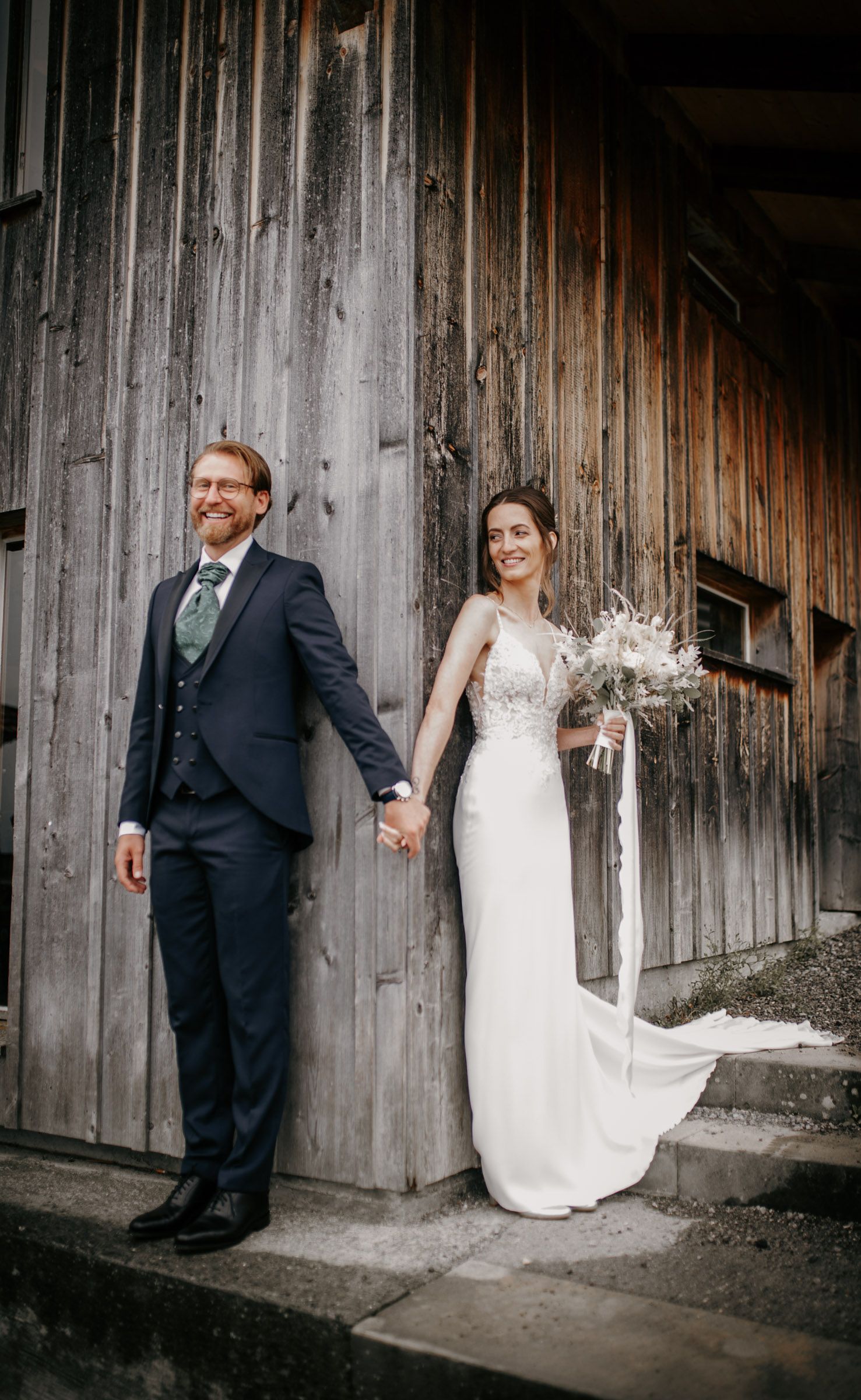 Bride and groom holding hands standing against a rustic wooden barn wall on their wedding day.