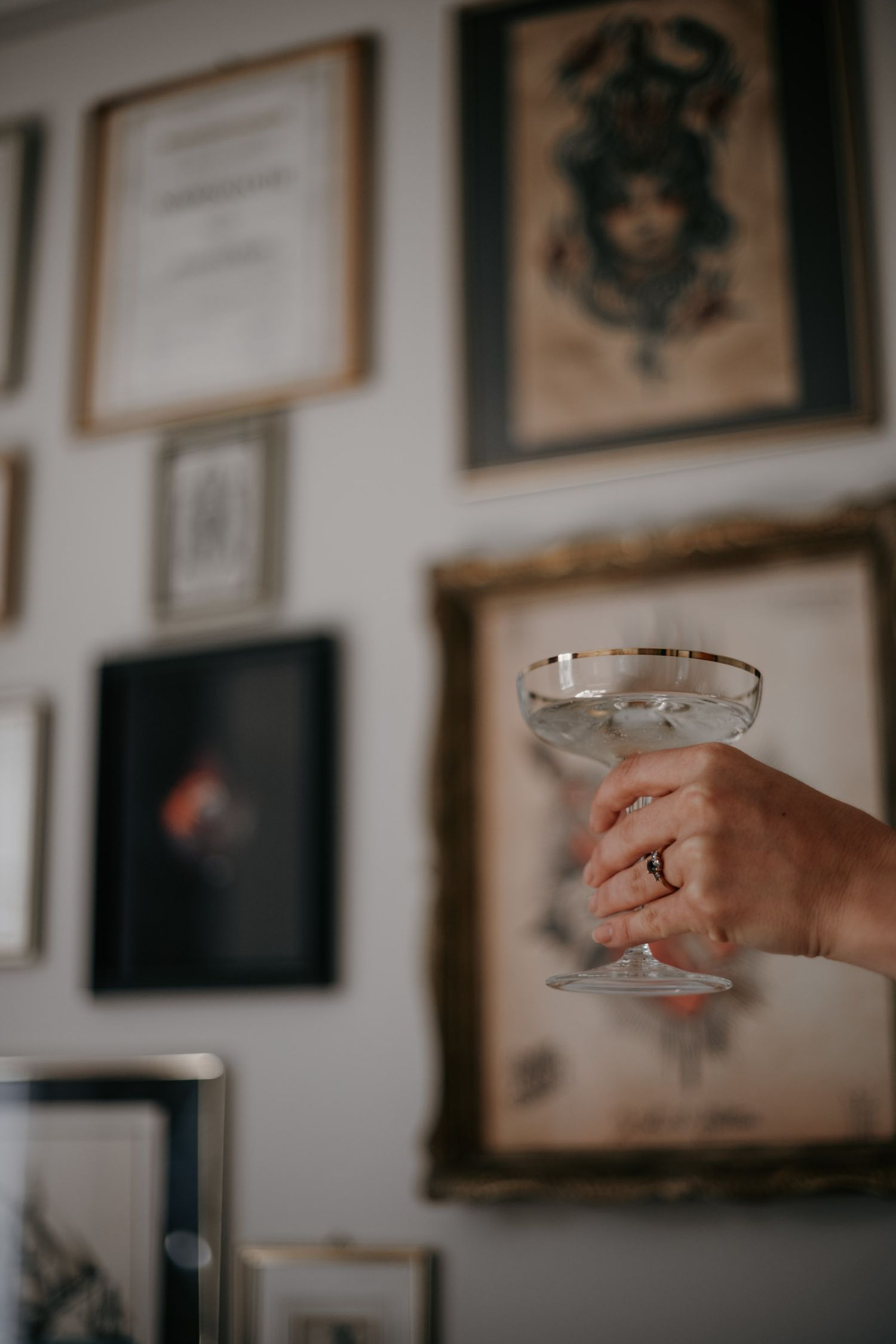Hand holding a vintage coupe glass with gold rim in front of a gallery wall with framed artwork.