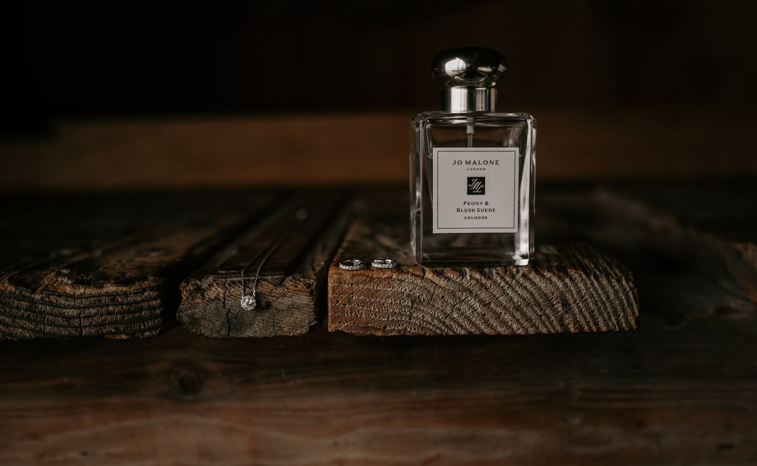 Jo Malone perfume bottle on rustic wooden surface with wedding rings in soft dark lighting.