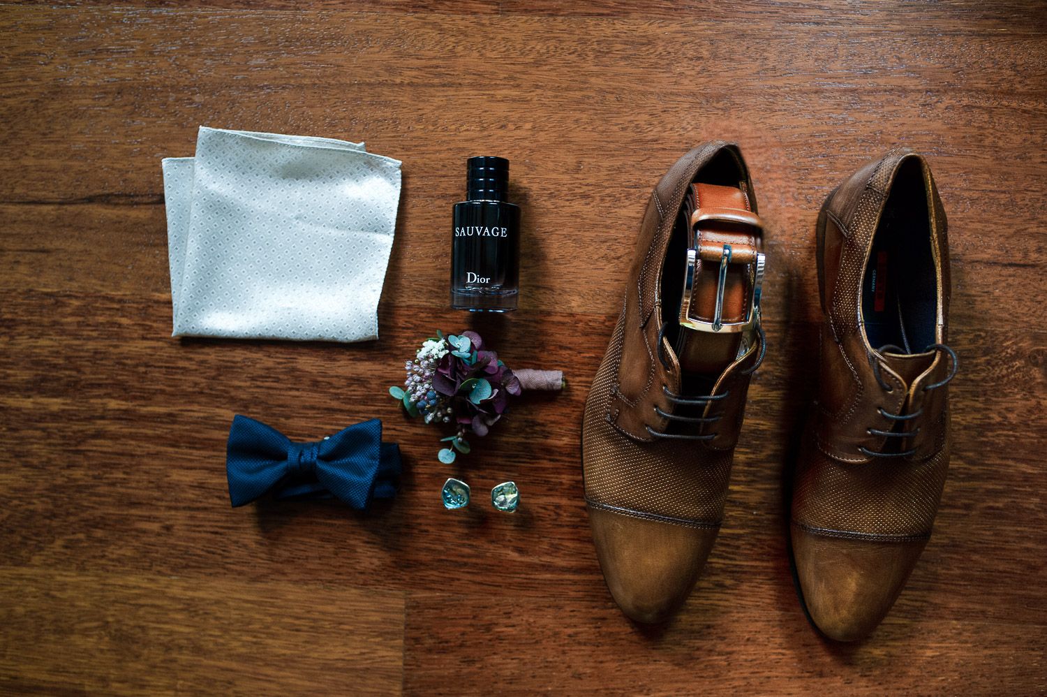 Groom accessories flat lay featuring brown oxford shoes, blue bow tie, cufflinks, and Dior cologne.