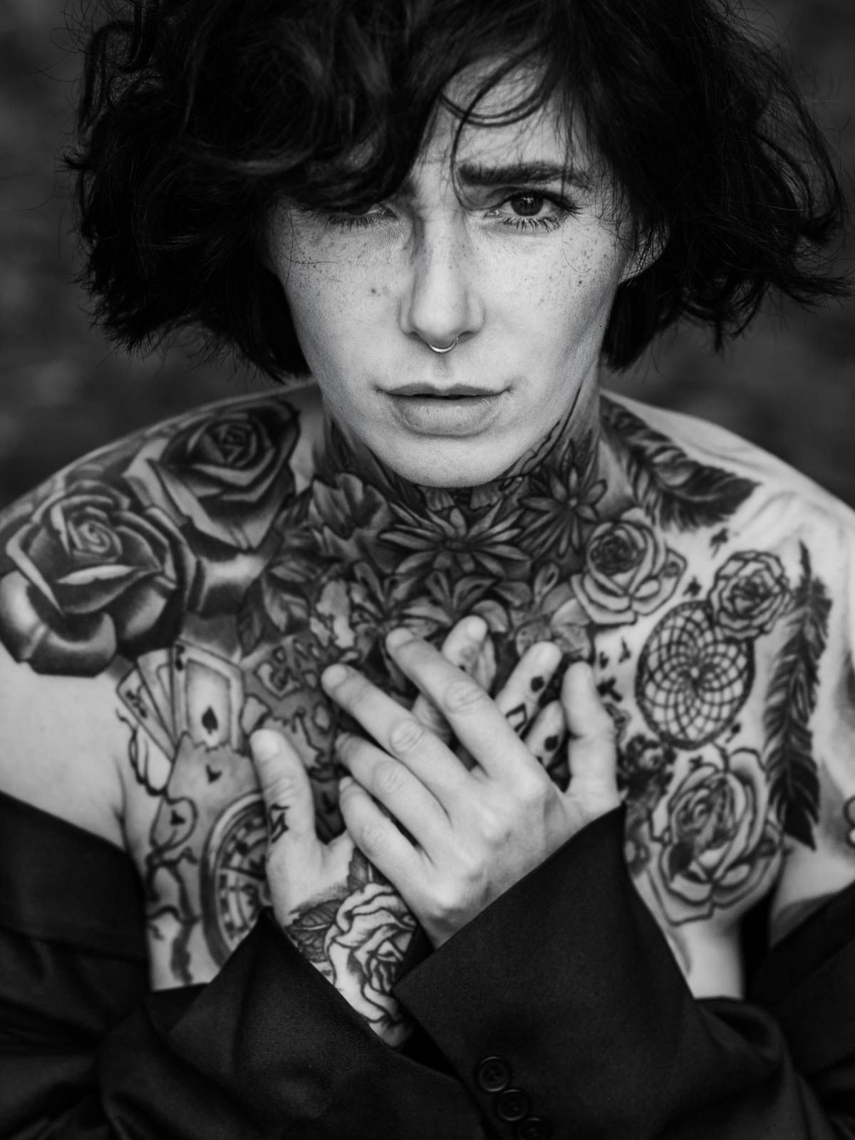 Black and white portrait of young woman with extensive floral chest and neck tattoos holding hands to chest.
