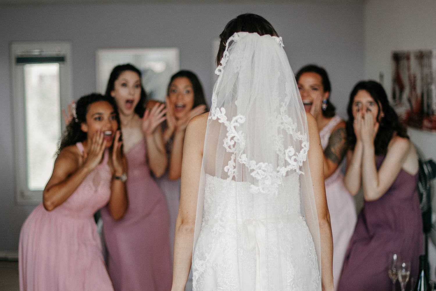 Bridesmaids in pink dresses reacting with surprise as bride in white gown and lace veil faces away.