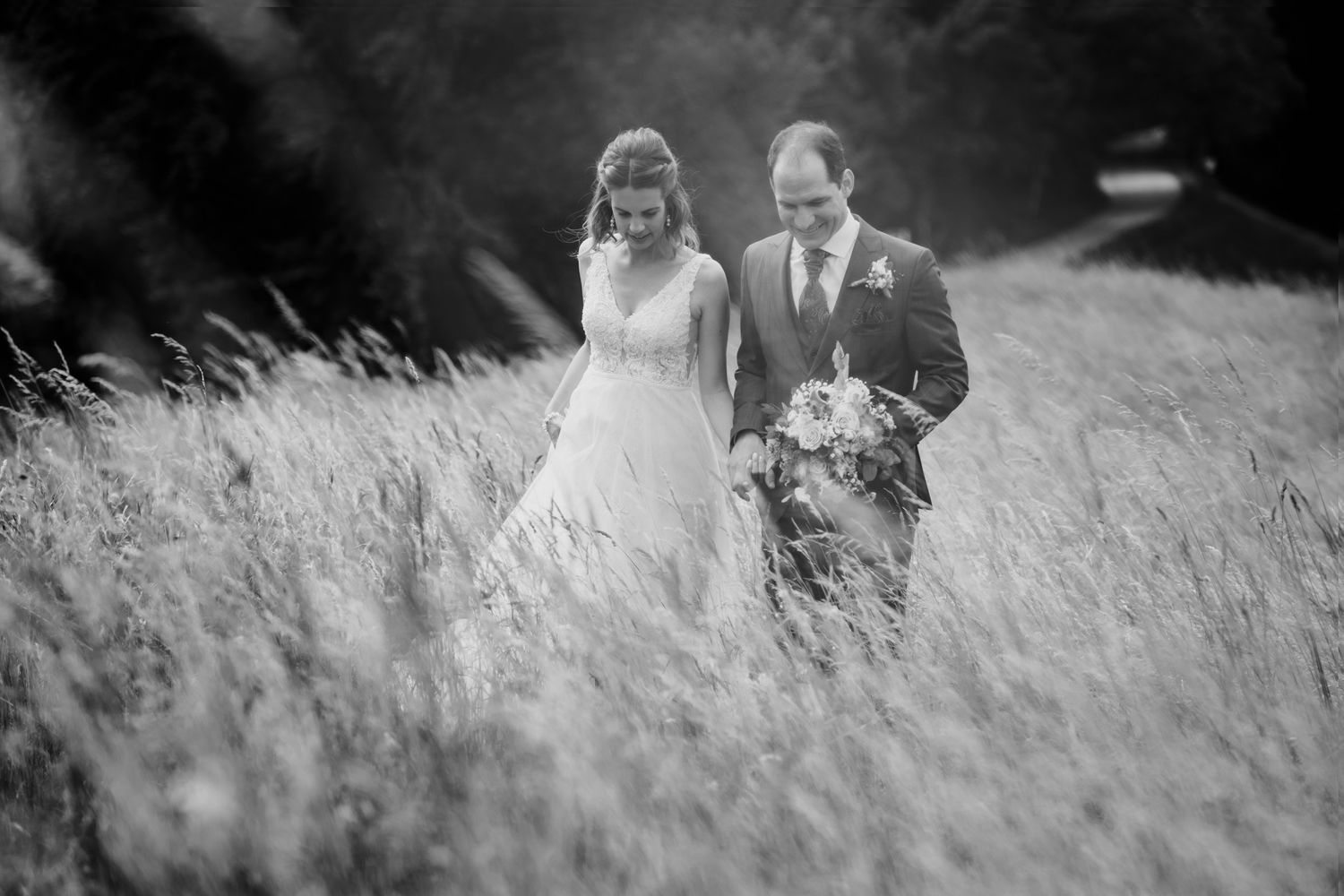 Black and white photo of bride and groom walking through a tall grass field on wedding day.