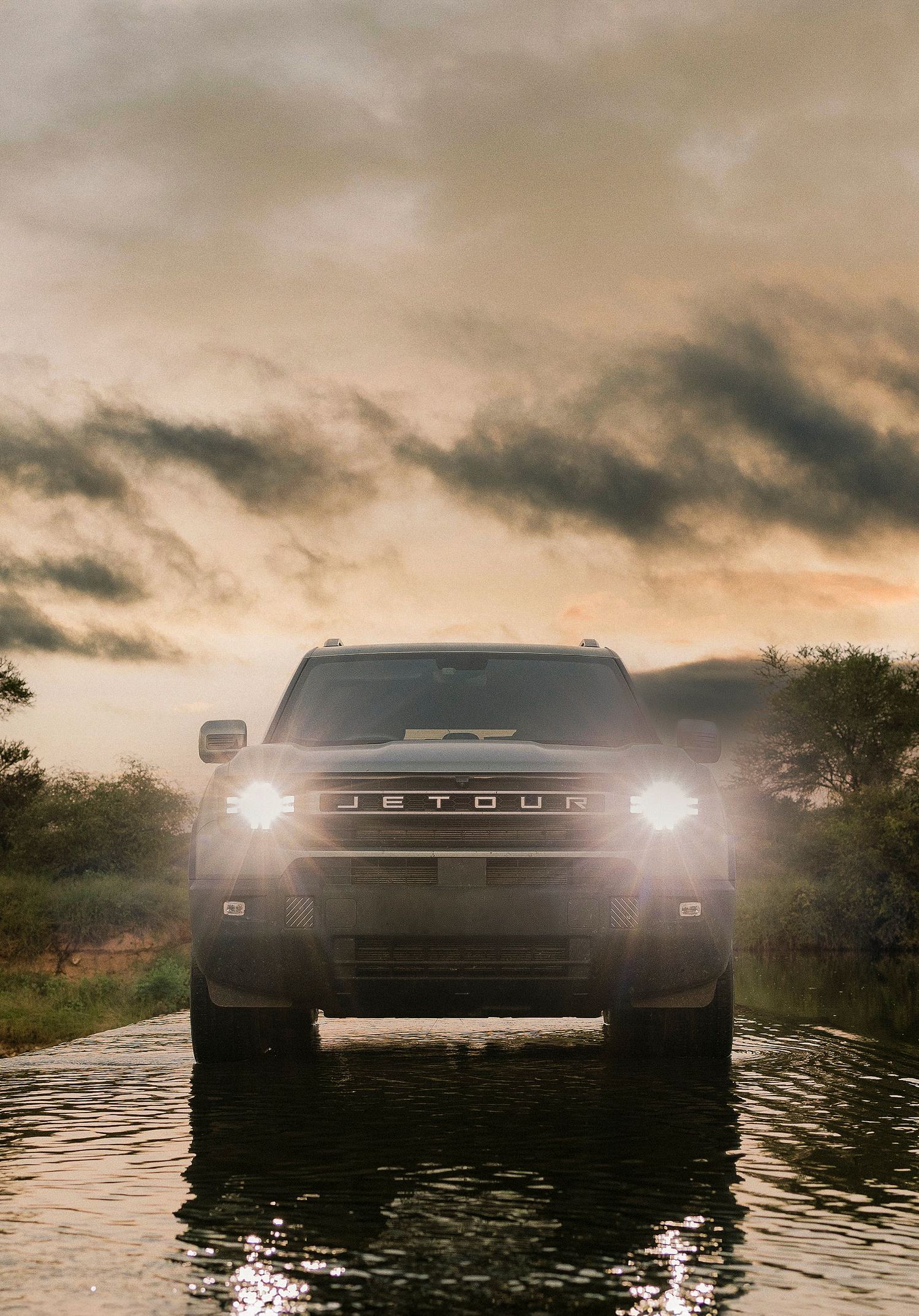 Jetour SUV with bright headlights driving through water at dramatic sunset with stormy clouds.