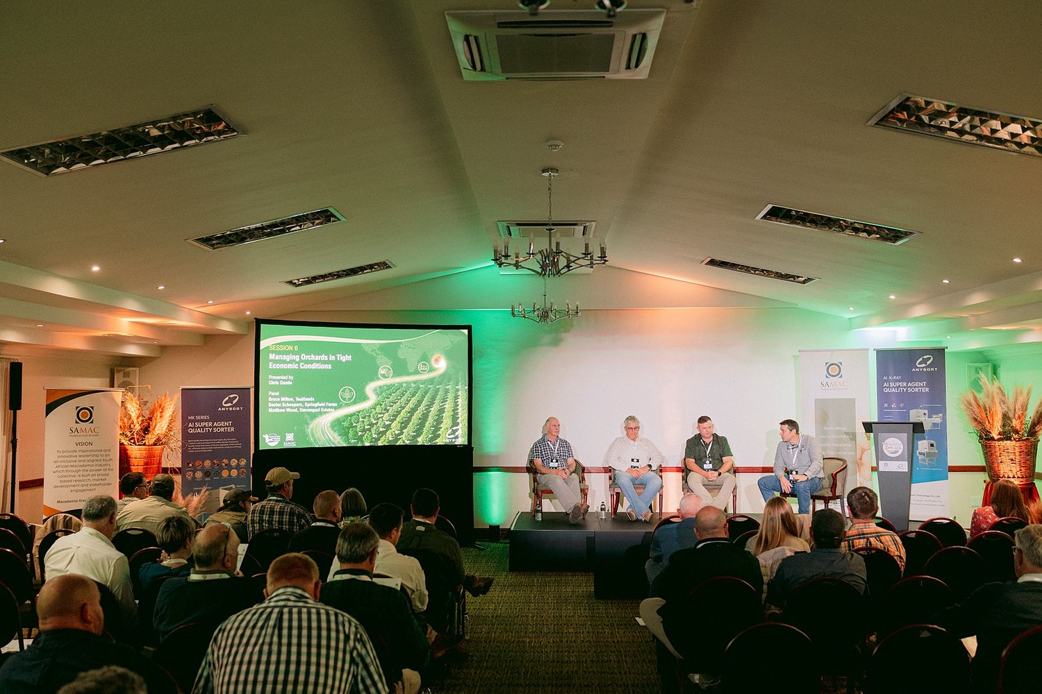 Audience seated in a conference room watches a panel of four speakers beside a large screen showing an aerial view of crop fields.