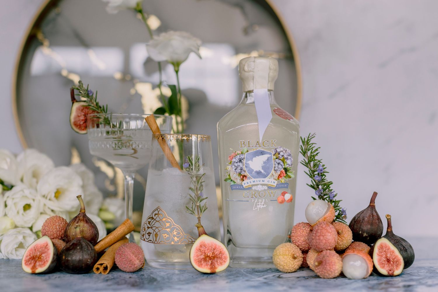 Elegant gin bottle display with fresh figs, lychees, rosemary, and glassware on a marble surface.
