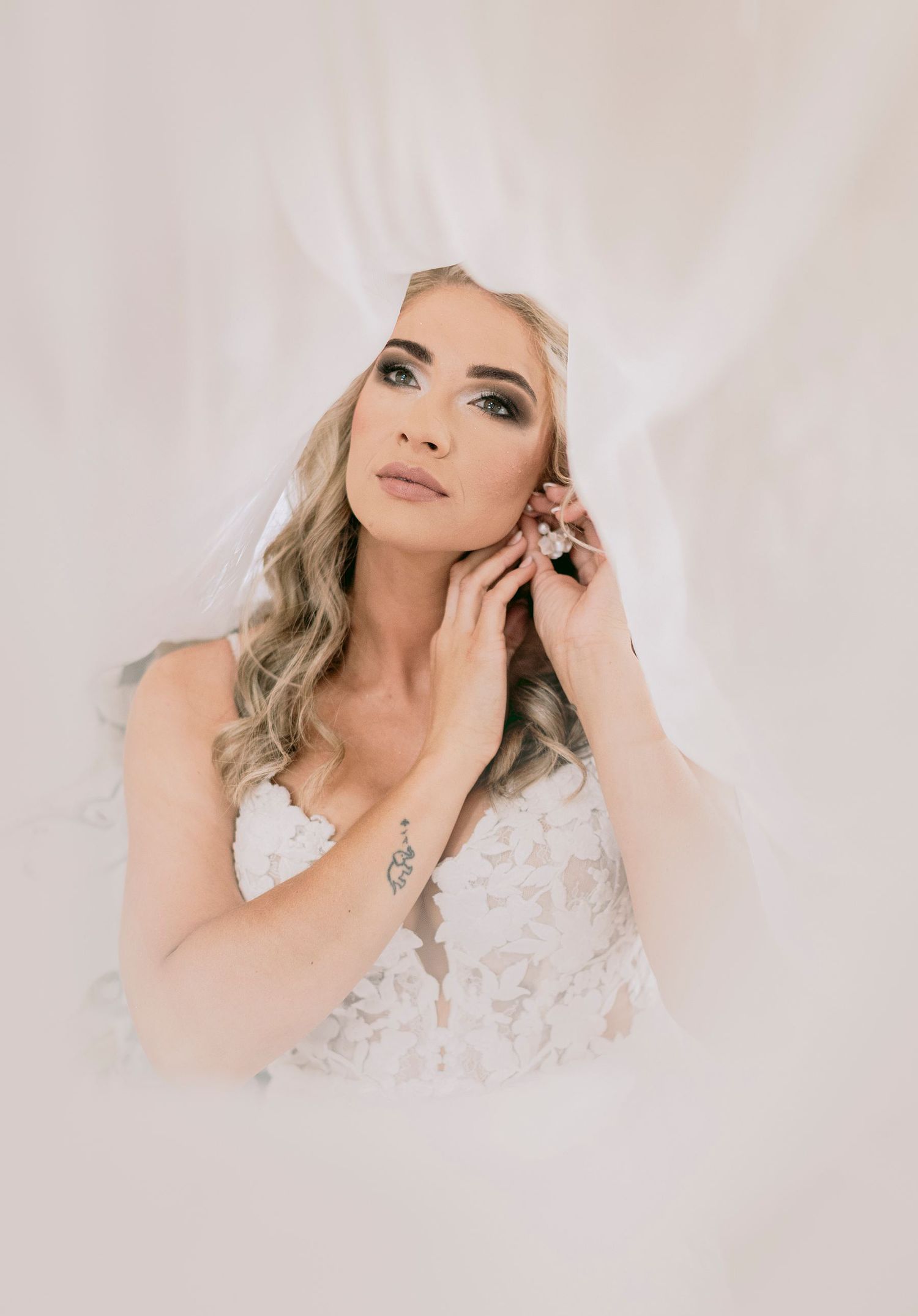 Beautiful bride in floral lace gown posing with hand to face against soft white fabric backdrop.