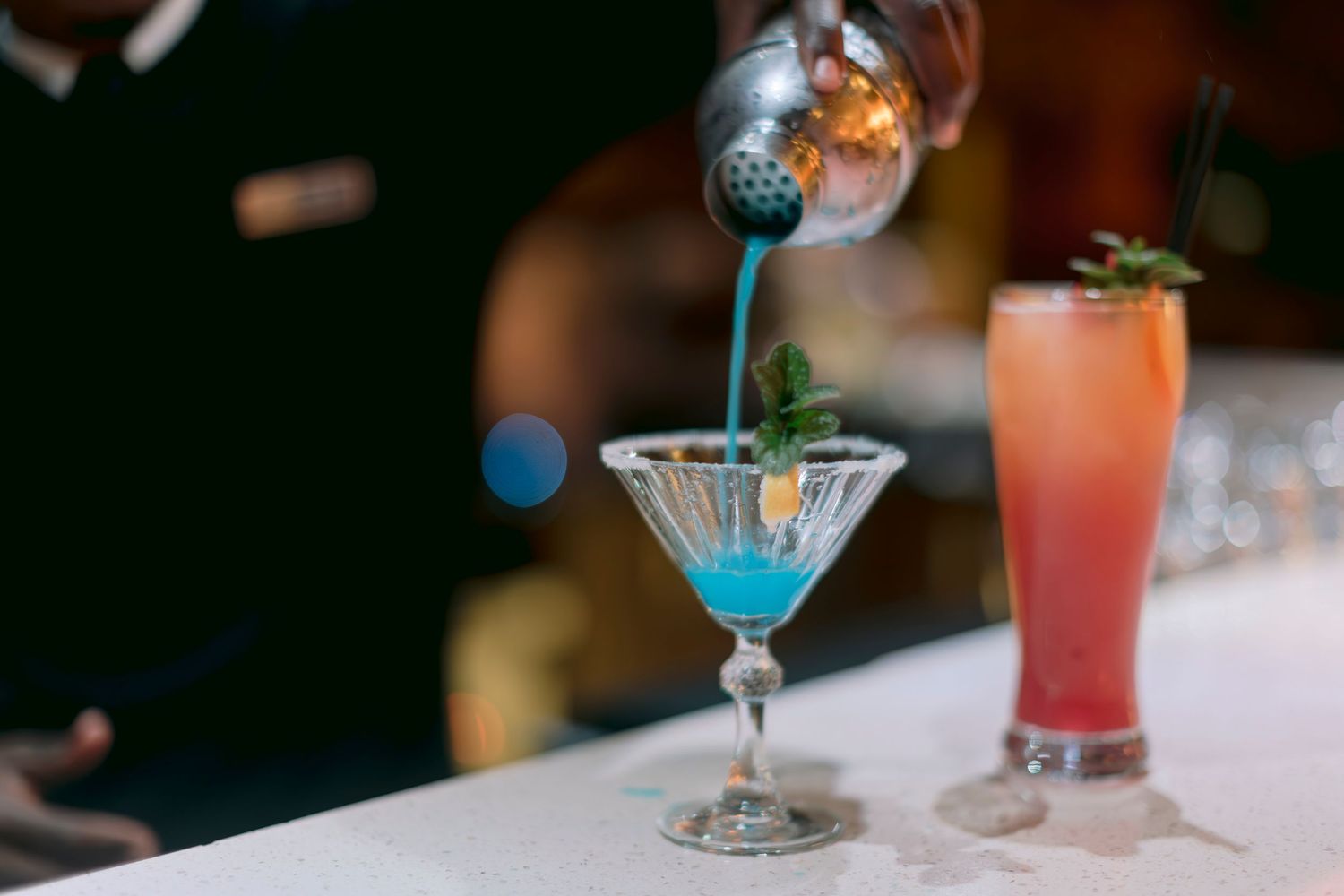 Bartender pouring a blue cocktail into a martini glass beside a red cocktail.