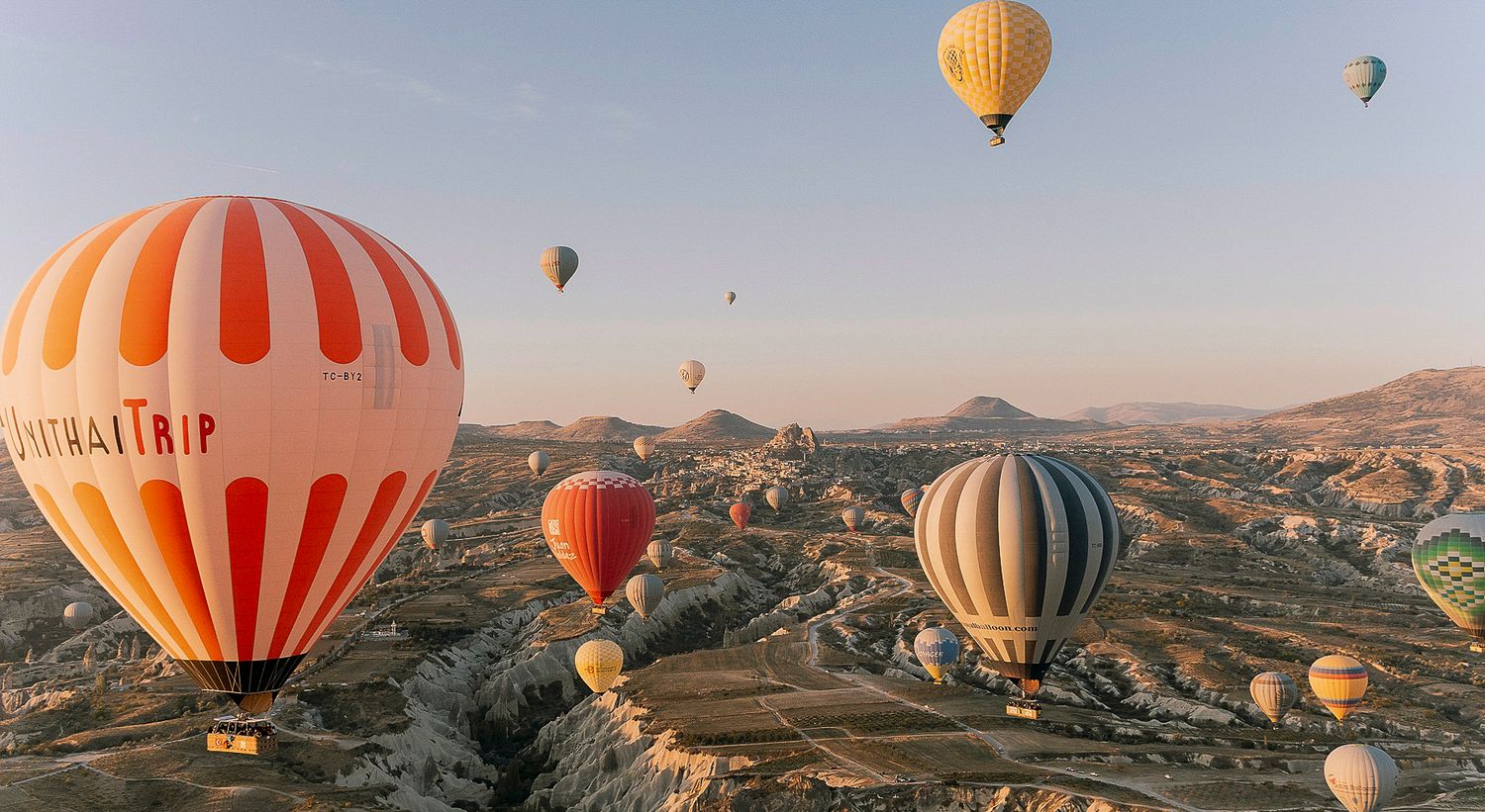 Colorful hot air balloons floating over rocky Cappadocia landscape at sunrise in Turkey.