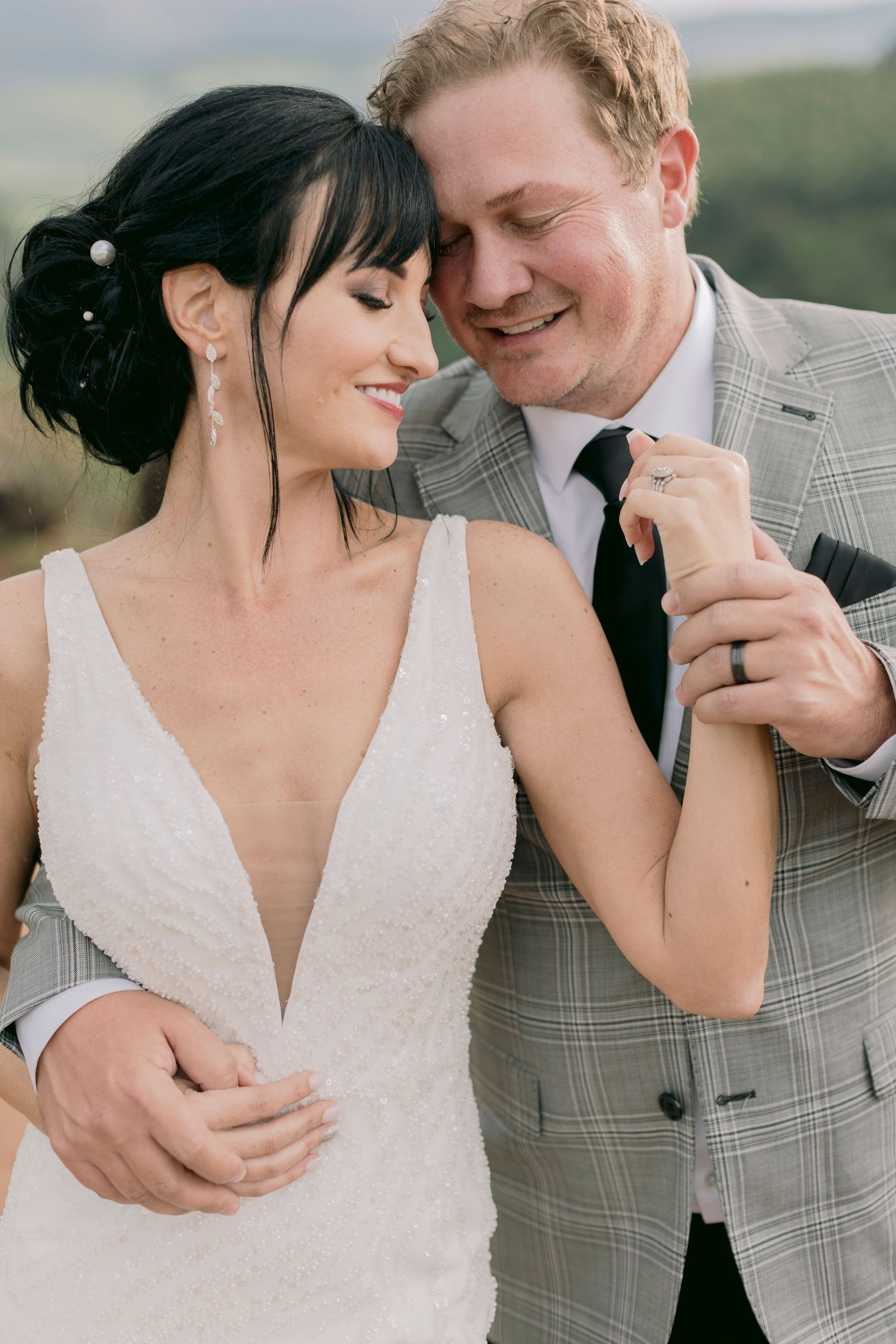Smiling bride in beaded V-neck gown and groom in plaid suit sharing tender moment outdoors.