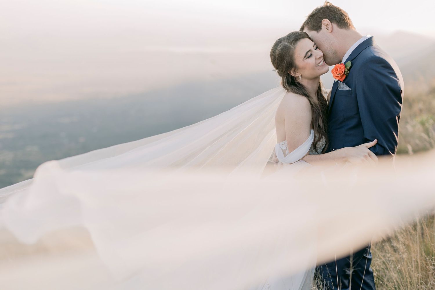 Romantic couple embracing on hilltop at sunset with flowing bridal veil billowing in the wind.
