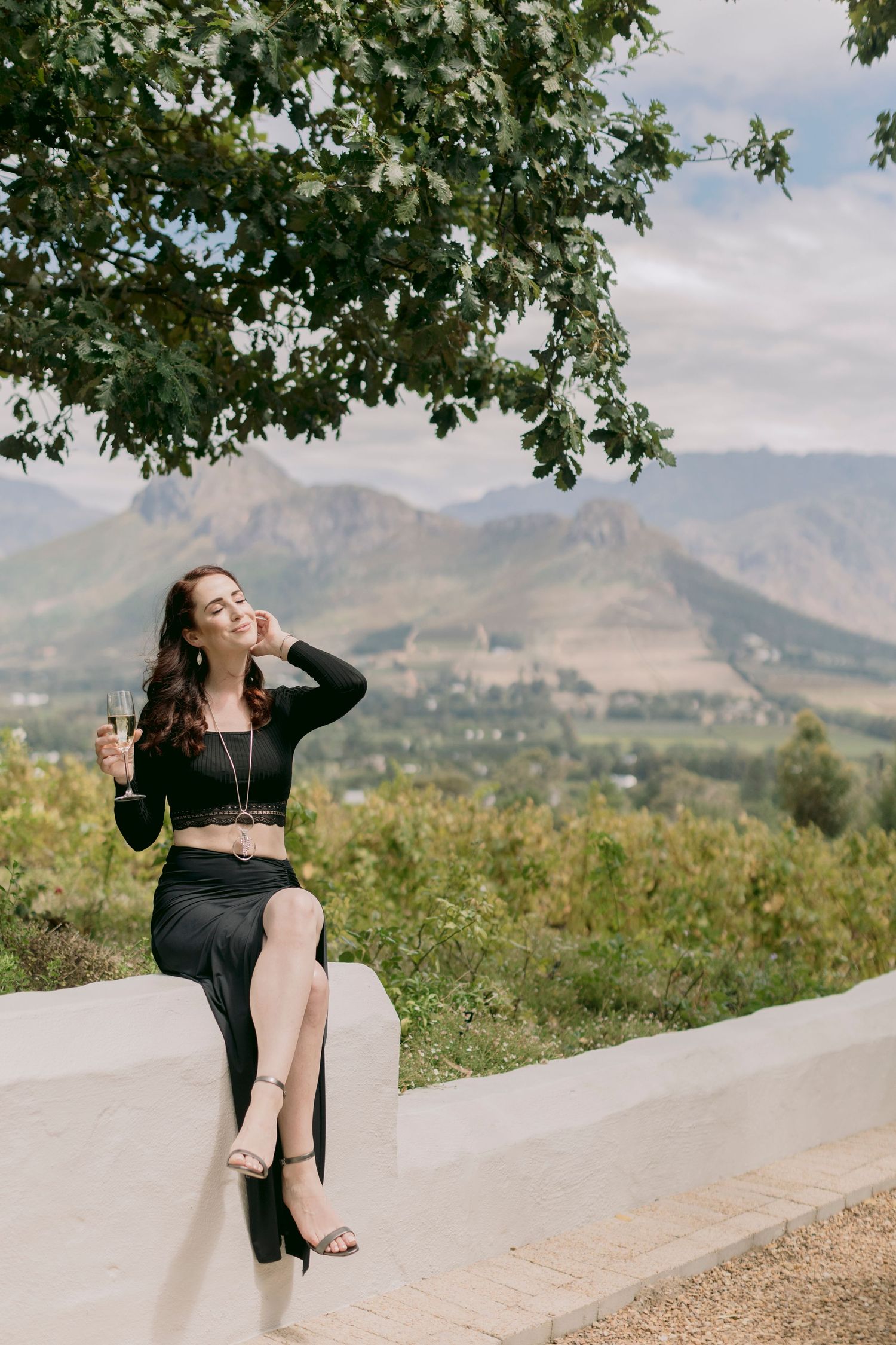 Elegant woman in black outfit sitting on a wall with mountain vineyard landscape behind her.