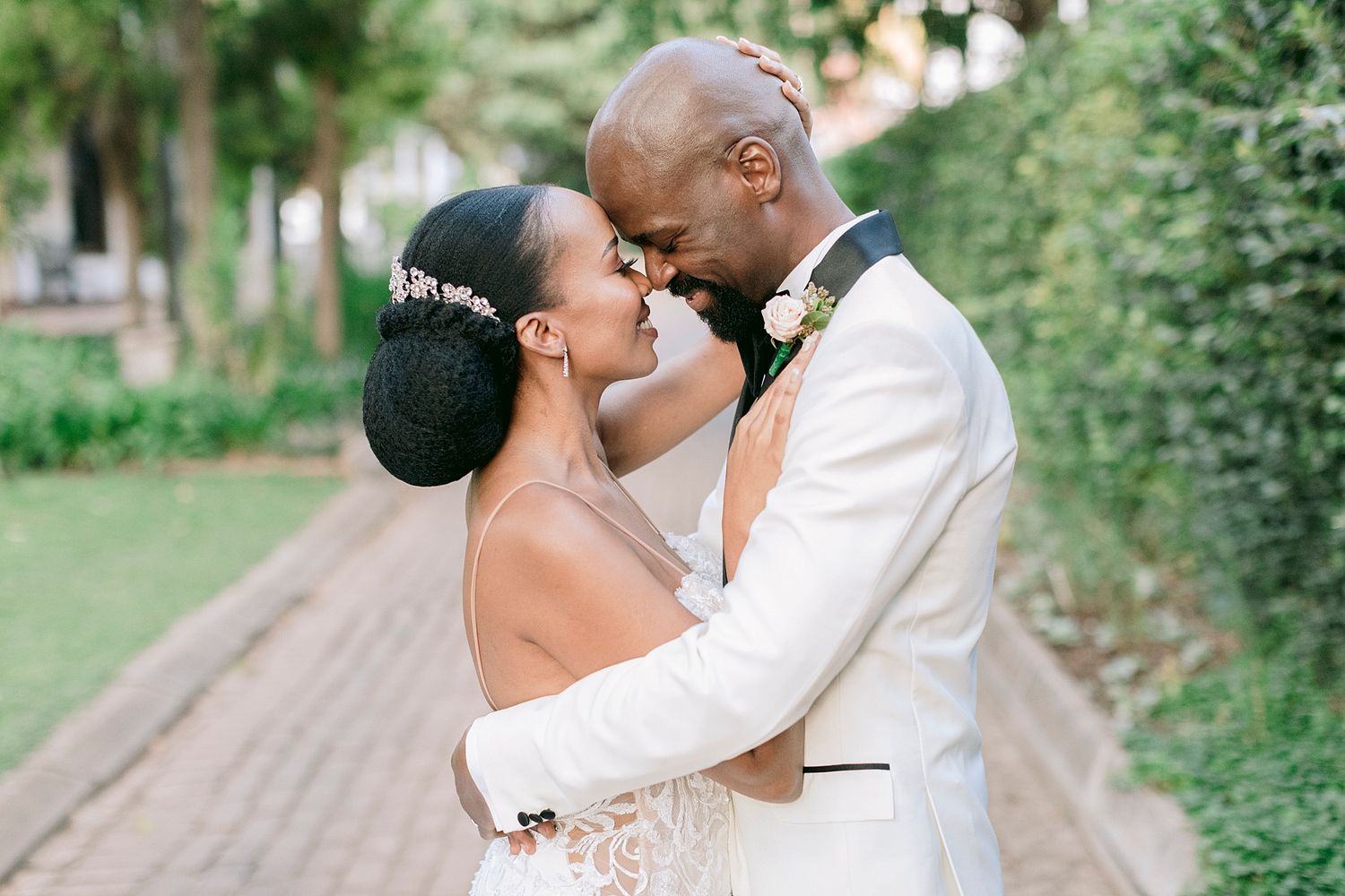 Bride and groom share an intimate moment nose-to-nose outdoors, bride in lace gown with floral hairpiece.