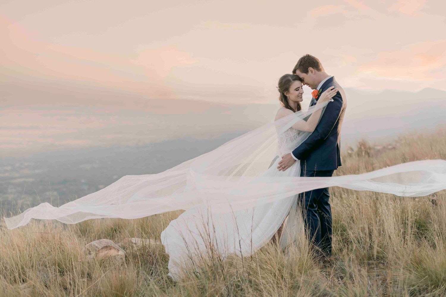 Bride and groom embracing on golden hillside at sunset with dramatic flowing cathedral veil.