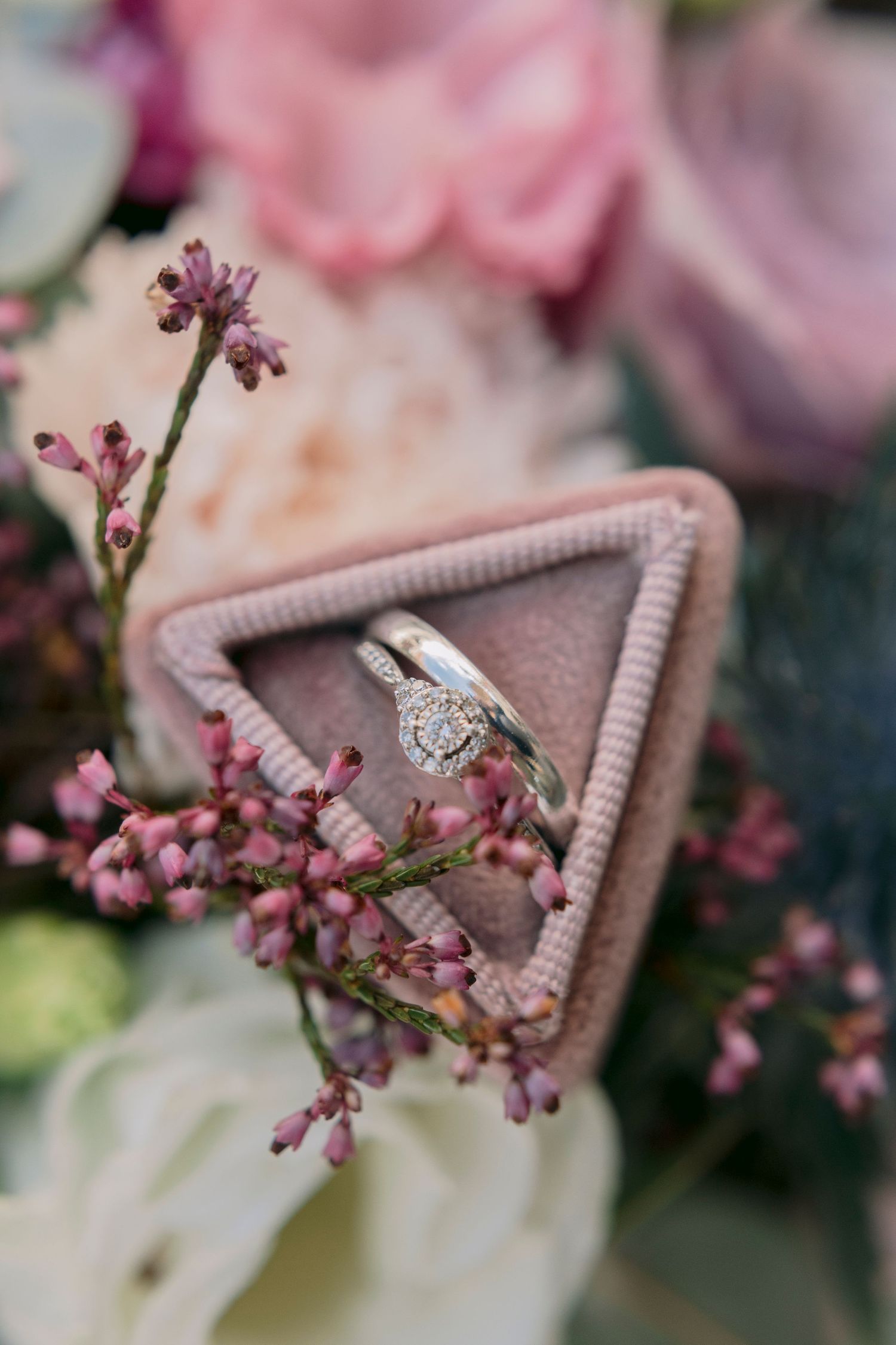 Diamond halo engagement ring and wedding band displayed in a pink velvet triangular ring box with flowers.
