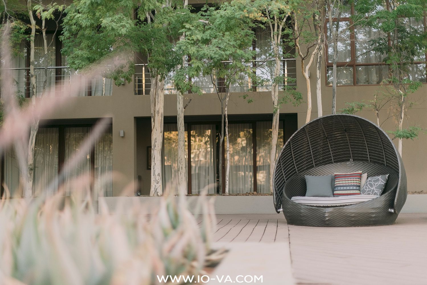 Woven wicker daybed pod with cushions on a wooden patio, framed by tall trees and a modern building with large glass windows.