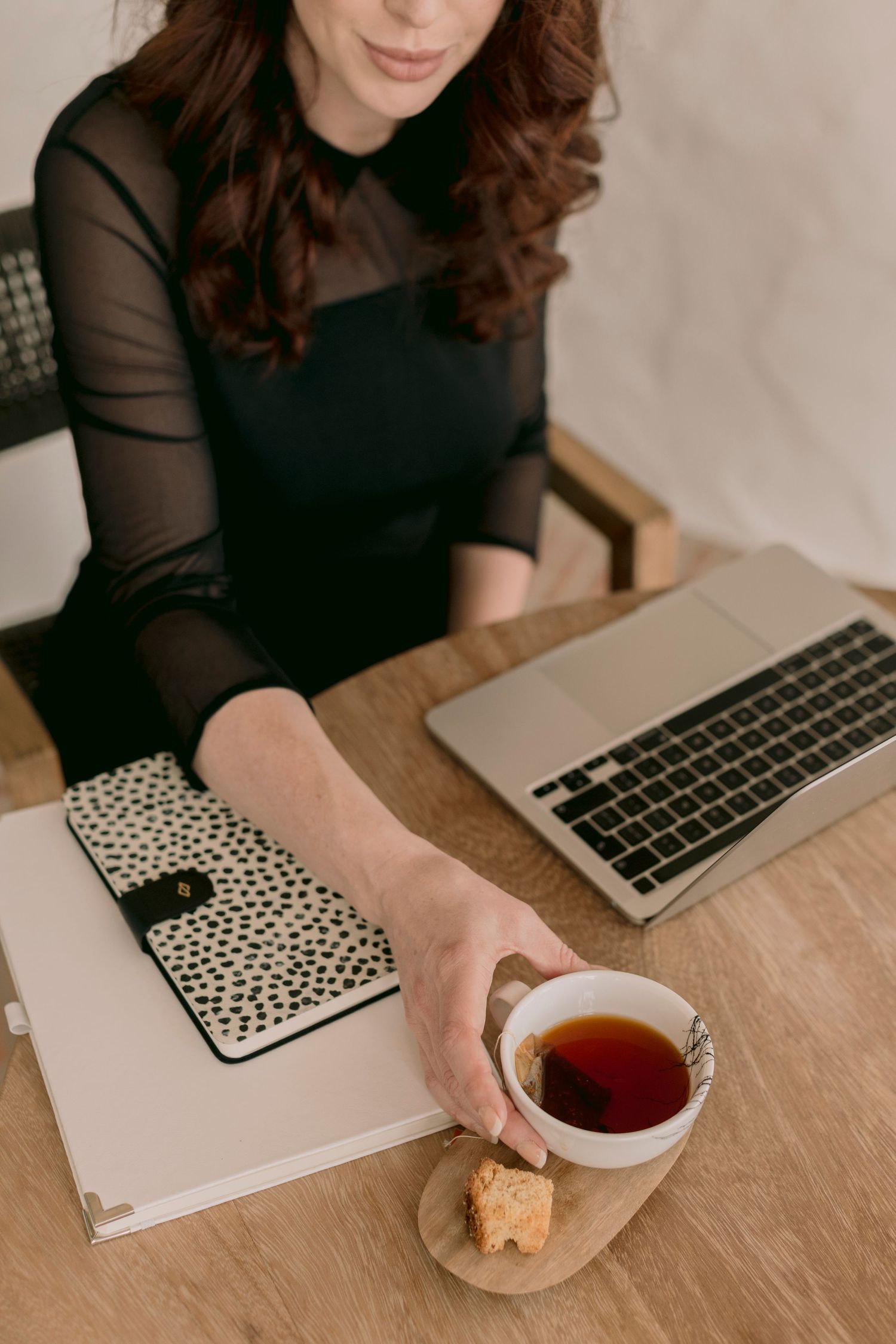 Woman in black dress reaching for tea cup beside laptop and spotted notebook on wooden desk.