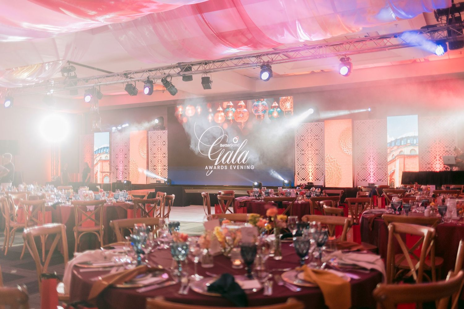 Elegant gala event venue with round tables, floral centerpieces, stage lighting, and large LED screen display.