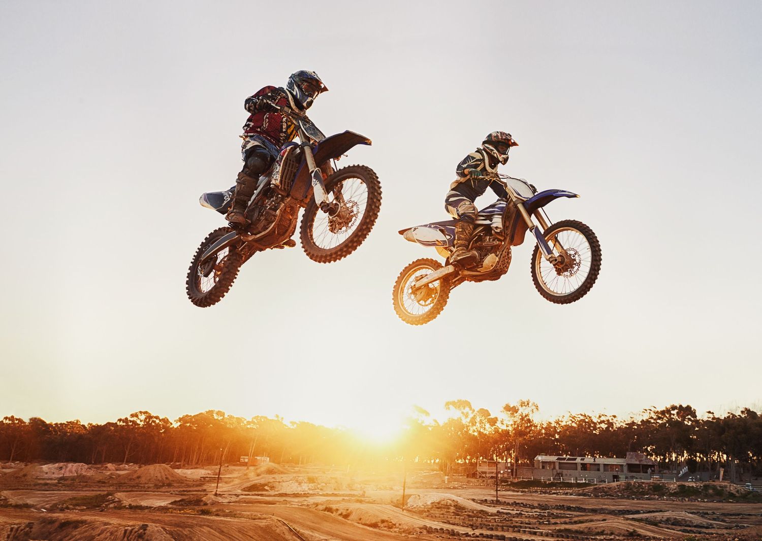Two motocross riders performing dramatic mid-air jumps on dirt bikes at sunset over an outdoor track.