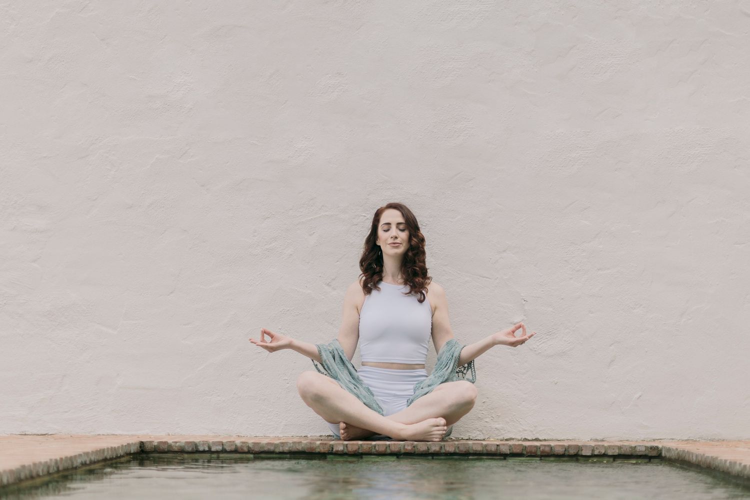 Young woman meditating in lotus pose by a pool against a white wall outdoors.