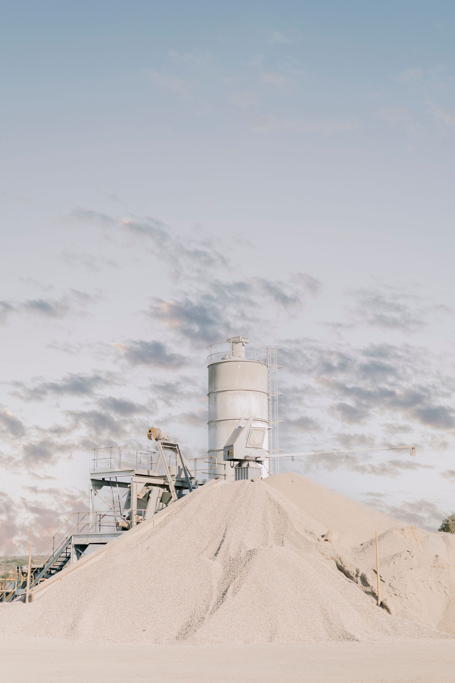 Industrial cement silo and conveyor equipment behind large pile of sand at dusk.