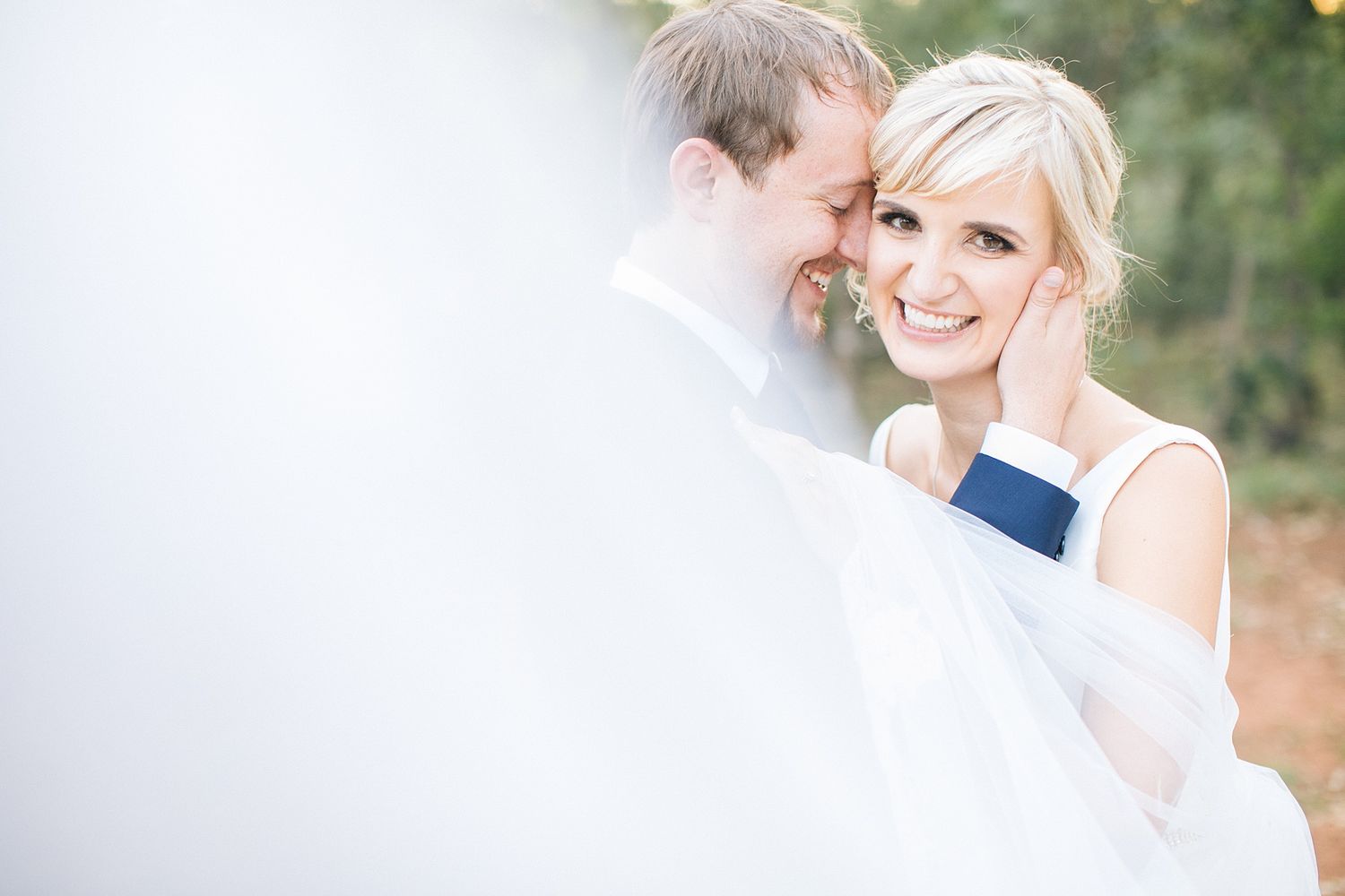A smiling bride in a white gown is tenderly embraced by her groom in a suit with a navy tie on their wedding day.