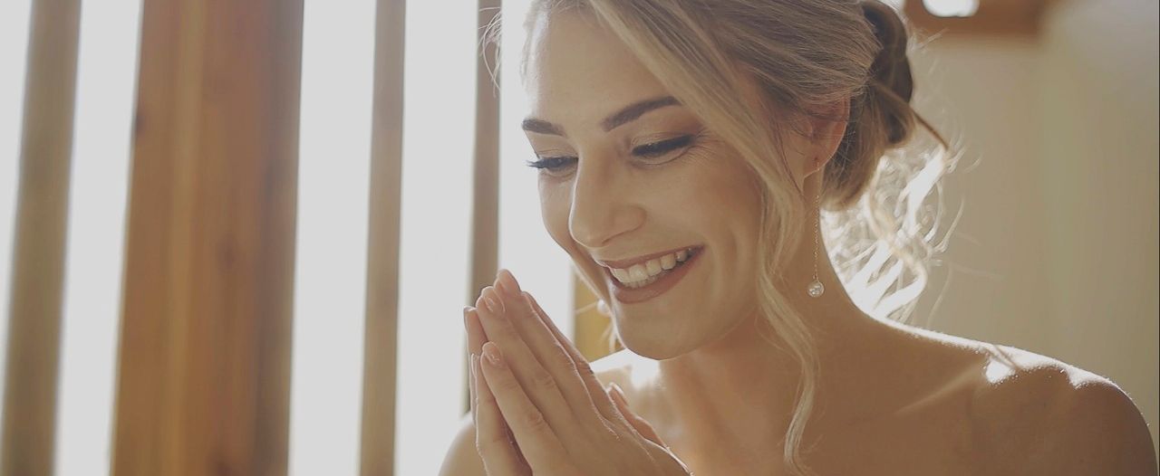 Radiant bride smiling with hands clasped near a sunlit wooden staircase railing.
