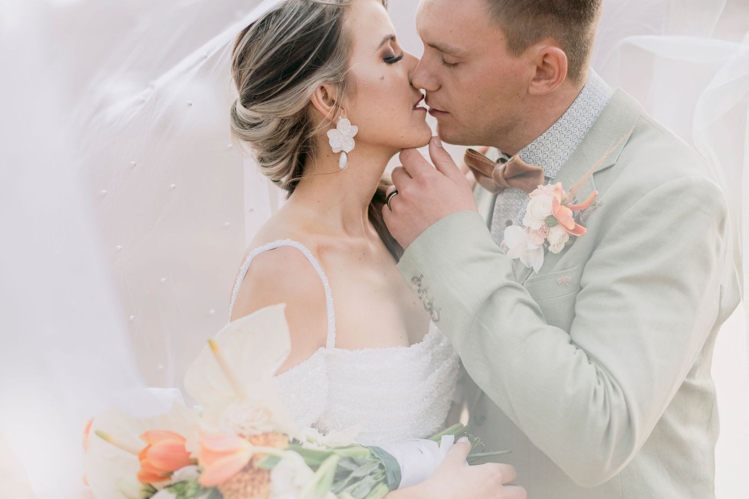Bride and groom sharing a kiss under veil with peach floral bouquet and soft romantic lighting.
